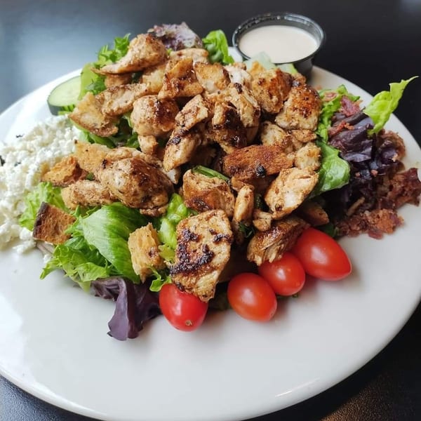 A grilled chicken salad with lettuce, tomatoes, feta, bacon, croutons, and a side of dressing.
